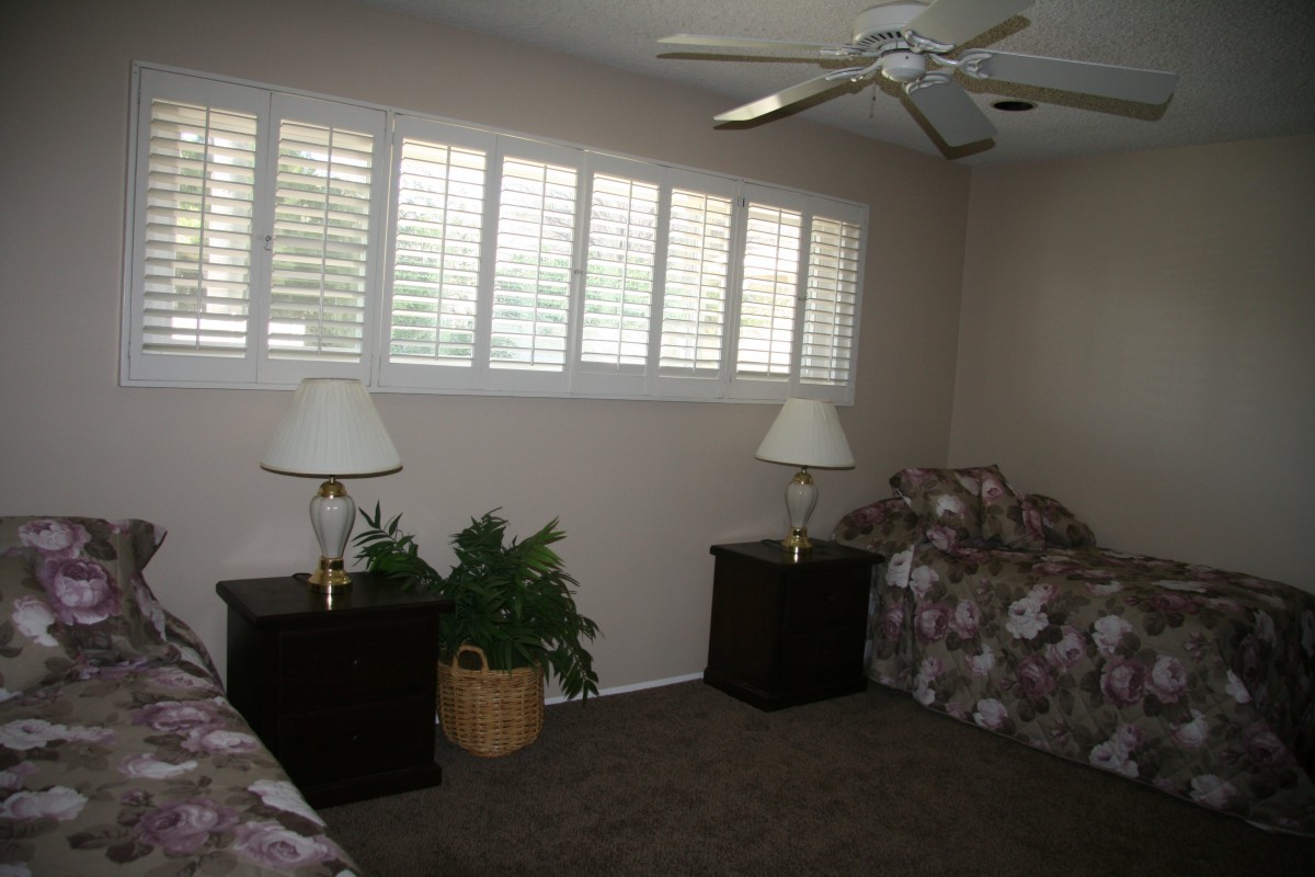 A bedroom interior featuring two twin beds with matching bedding, nightstands with lamps, a ceiling fan, and window shutters.