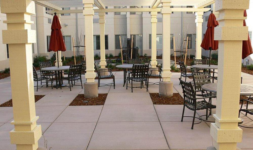 An outdoor patio with tables and chairs under a pergola, featuring red umbrellas.