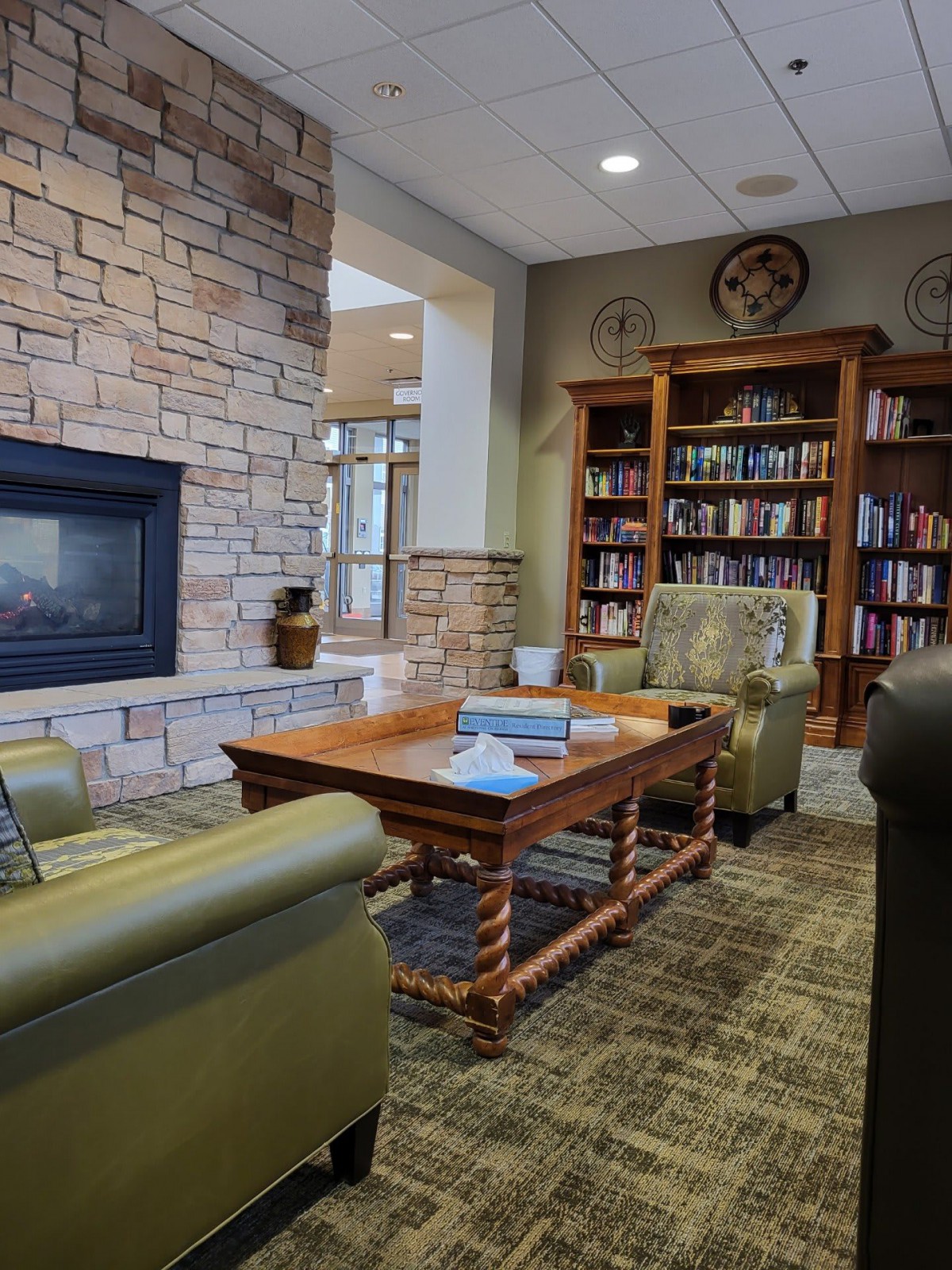 A cozy library corner with comfortable armchairs, a wooden coffee table, and a fireplace set against bookshelves filled with books.