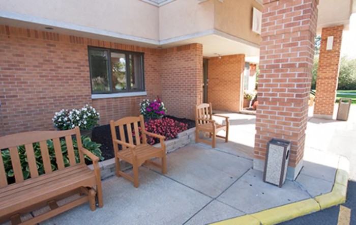 A small outdoor seating area with wooden chairs and flower beds next to the entrance of a brick building.