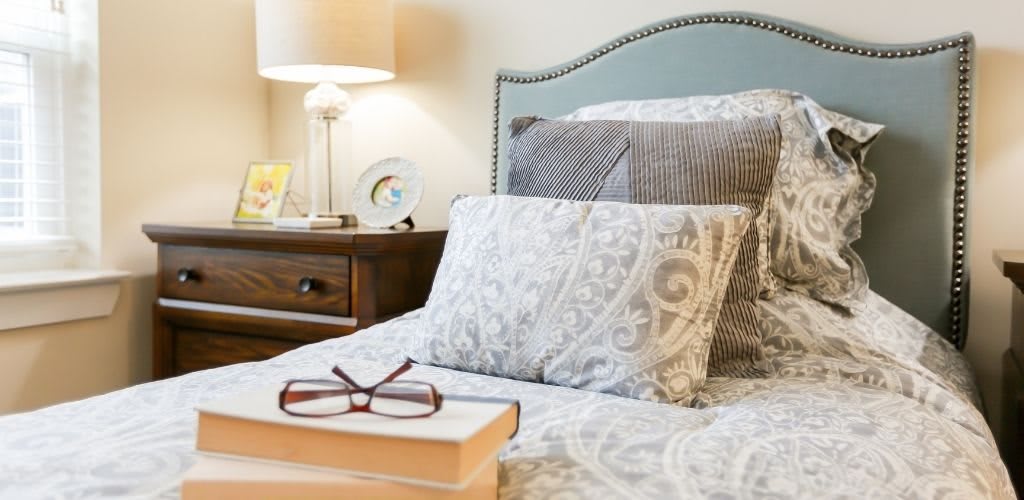A cozy bedroom setup with a neatly made bed, decorative pillows, and a nightstand featuring a lamp, glasses, and a book.