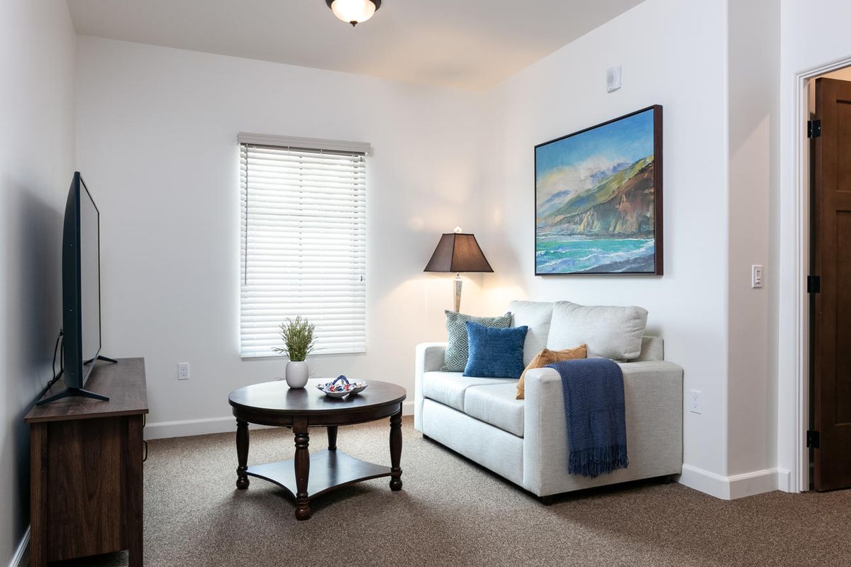 A cozy living room with a white sofa, a round coffee table, and a floor lamp, featuring artwork on the wall and natural light coming through the window.