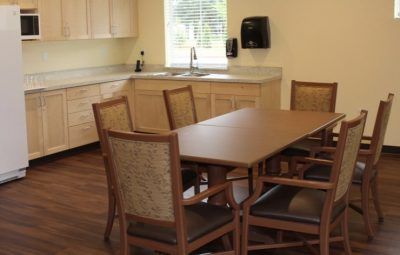 A clean and simple dining area with a table and chairs, adjacent to a kitchen, featuring wooden floors and natural lighting from a window.