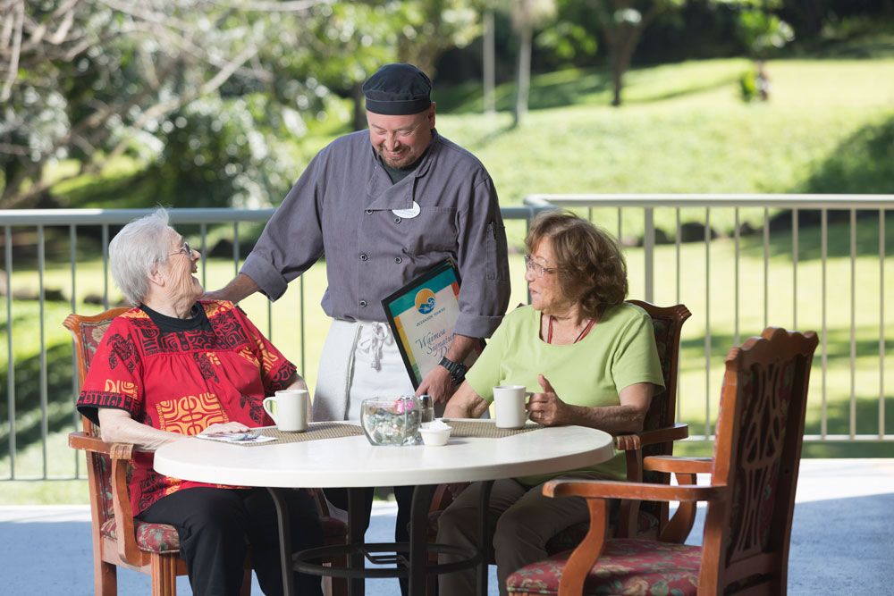 A waiter attending to two elderly women dining outdoors on a sunny day.