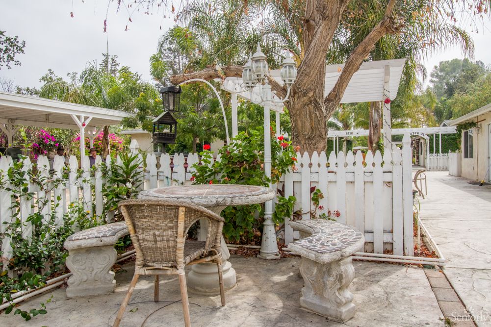 A charming garden patio with a wicker chair, table, white picket fence, and lush greenery.