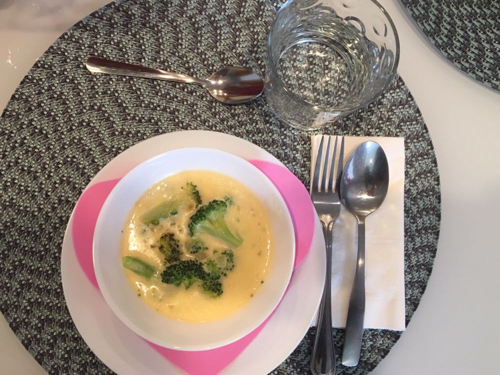 A bowl of soup with broccoli served on a table alongside utensils and a glass.