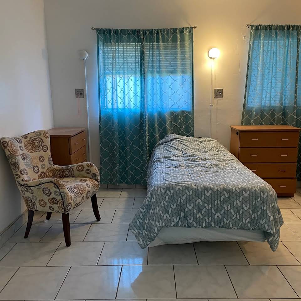 A simple bedroom with a single bed, armchair, dresser, and blue curtains.