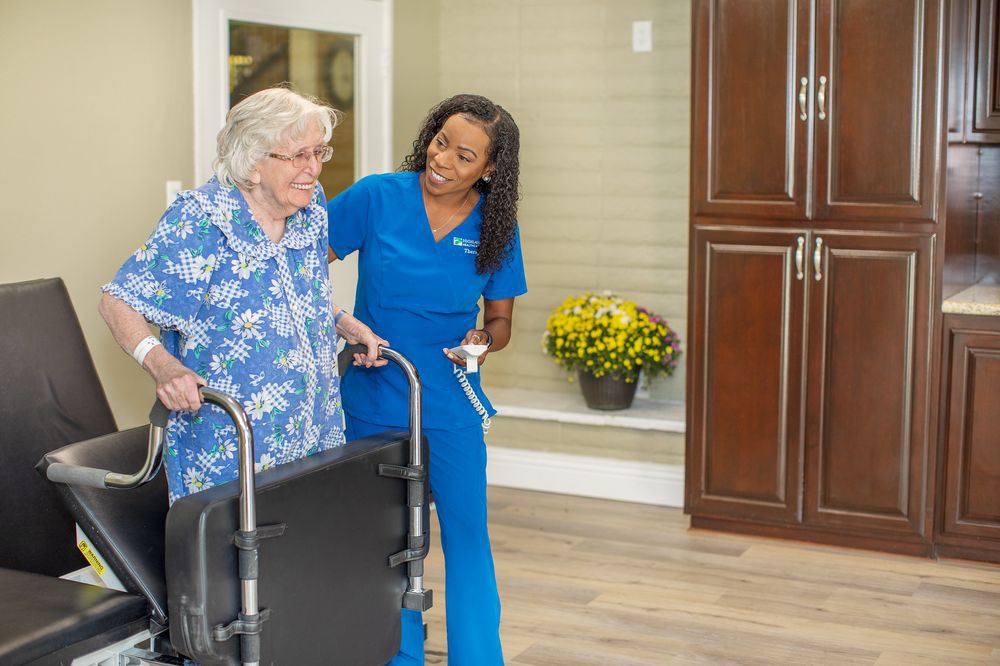 A caregiver assists a senior woman using a walker in a nursing home setting.