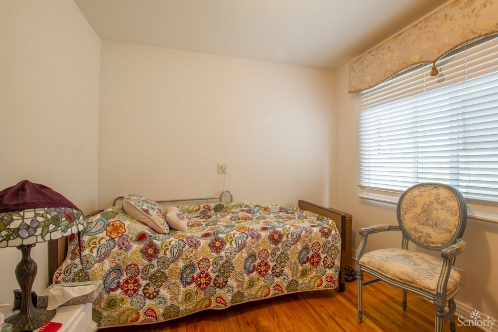 A cozy bedroom with a single bed covered in colorful bedding, an armchair by the window, and hardwood flooring.