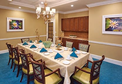 An elegant dining room with a large table set for a formal meal, featuring blue napkins, cushioned chairs, and a chandelier overhead.