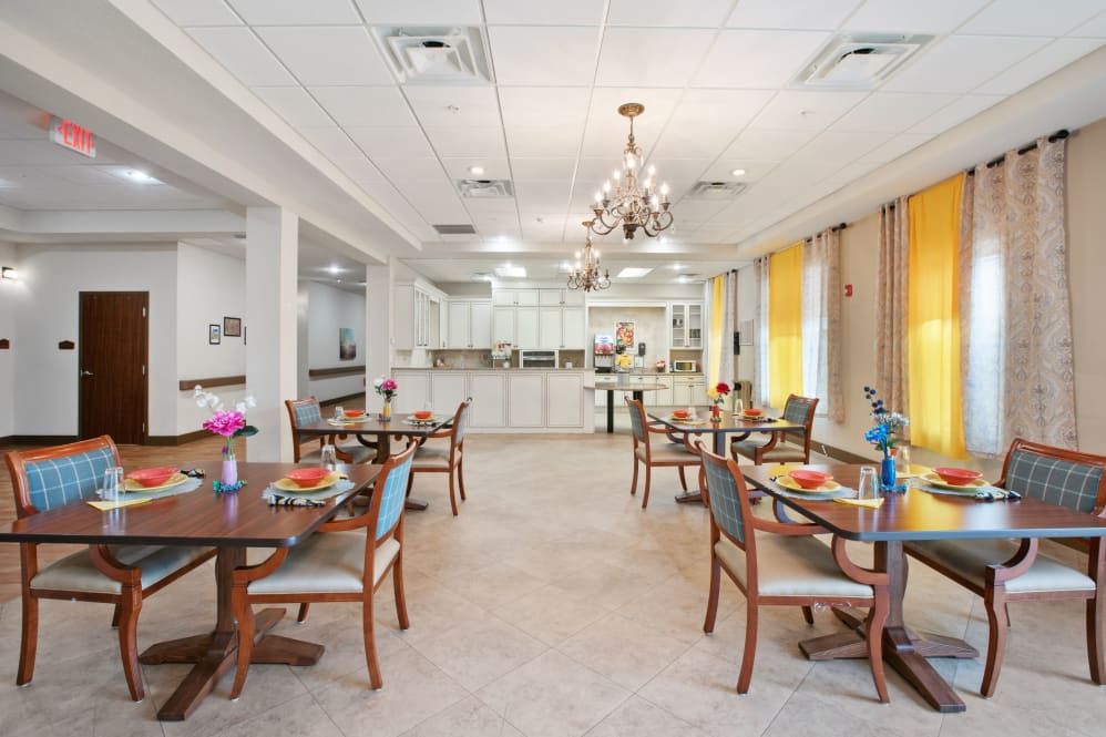 A well-lit dining area with elegantly arranged tables and chairs, featuring a chandelier and decorative flowers.