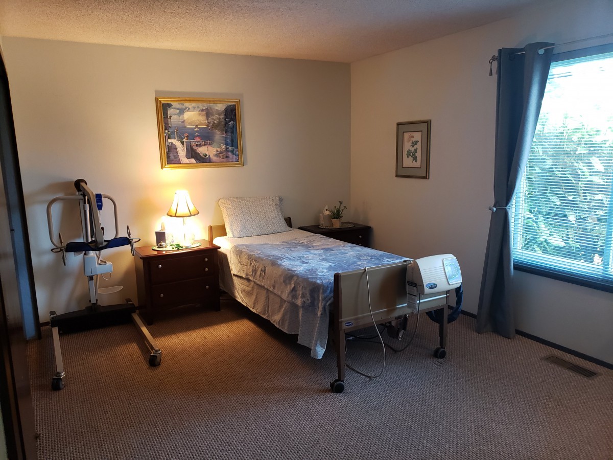 A cozy bedroom with a hospital bed, warmly lit by a lamp on a nightstand, and decorated with a painting on the wall.