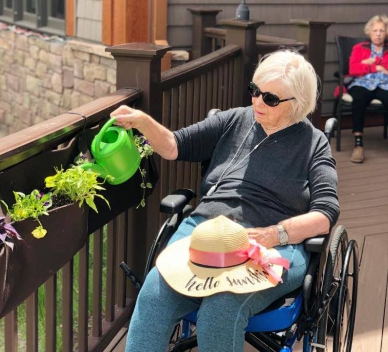 An elderly person in a wheelchair is watering plants outside, wearing sunglasses and holding a sun hat.