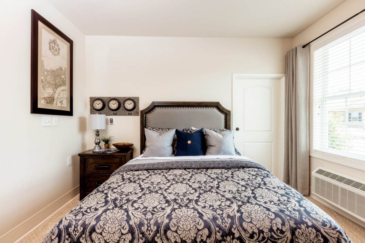 A traditionally styled bedroom featuring a bed with patterned bedding, flanked by a nightstand and decorated with wall art.