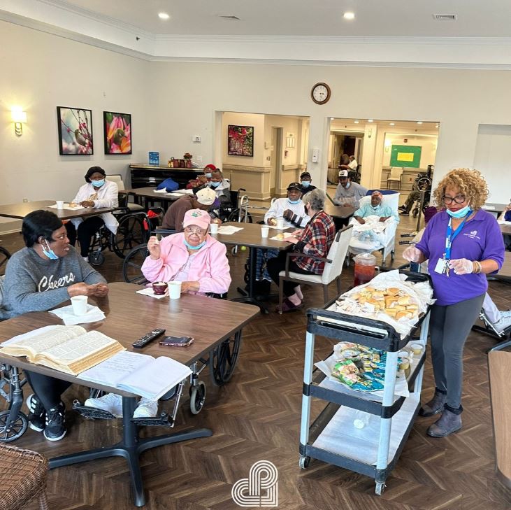 Residents socializing in a nursing home dining area with a healthcare worker serving meals.