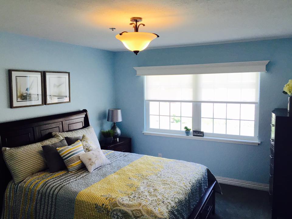 A cozy bedroom with blue walls, featuring a bed, nightstand, and large window.