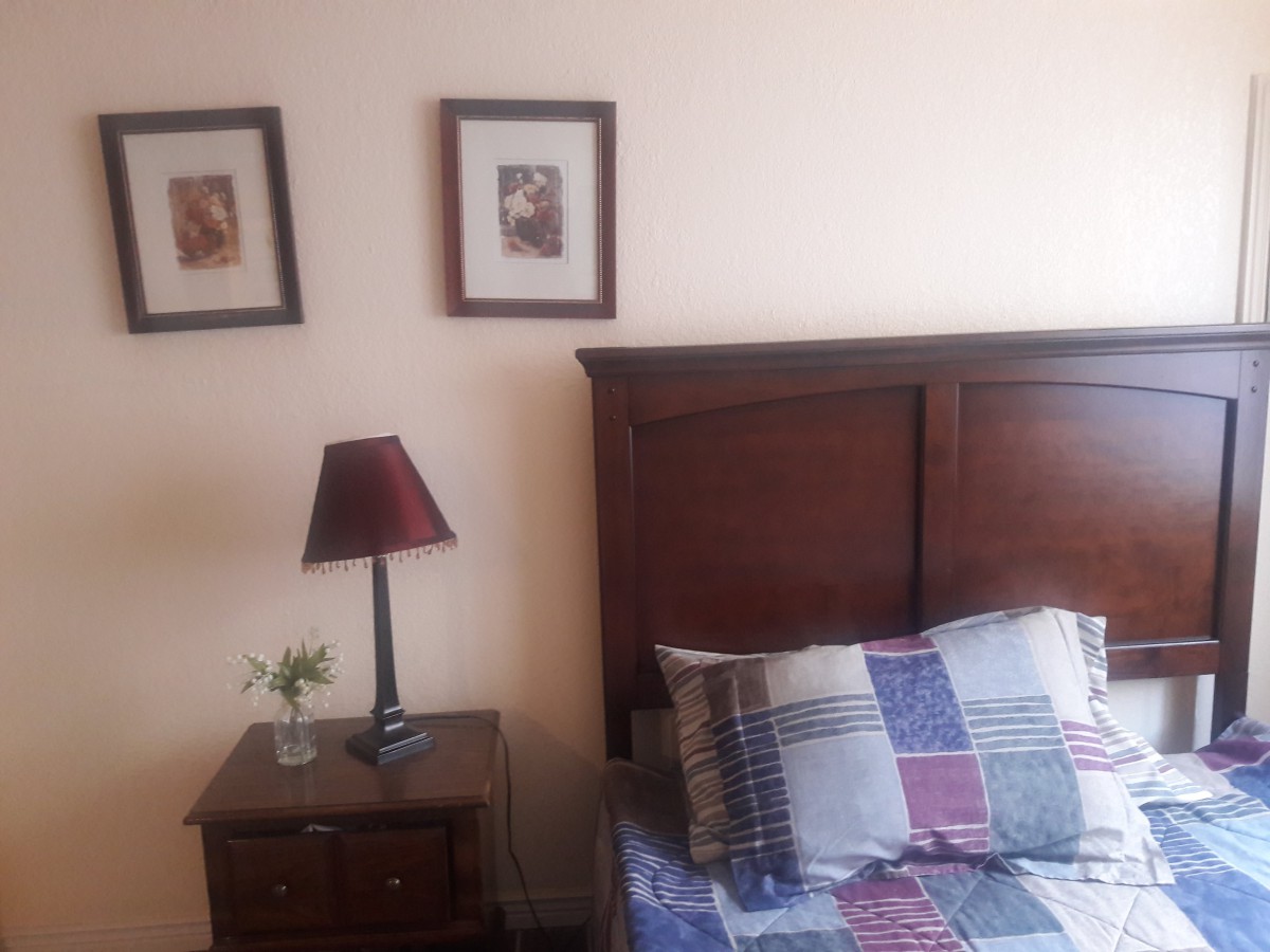 A cozy bedroom corner featuring a wooden bed, a quilted pillow, and a nightstand with a lamp and plant.