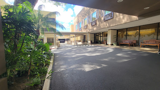 A serene courtyard view of a building, featuring palm trees and a spacious parking area illuminated by sunlight.