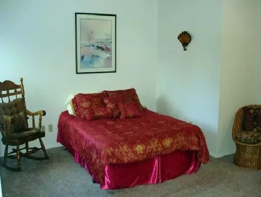 A cozy bedroom featuring a bed with red bedding, a wooden chair, and artwork on the wall.
