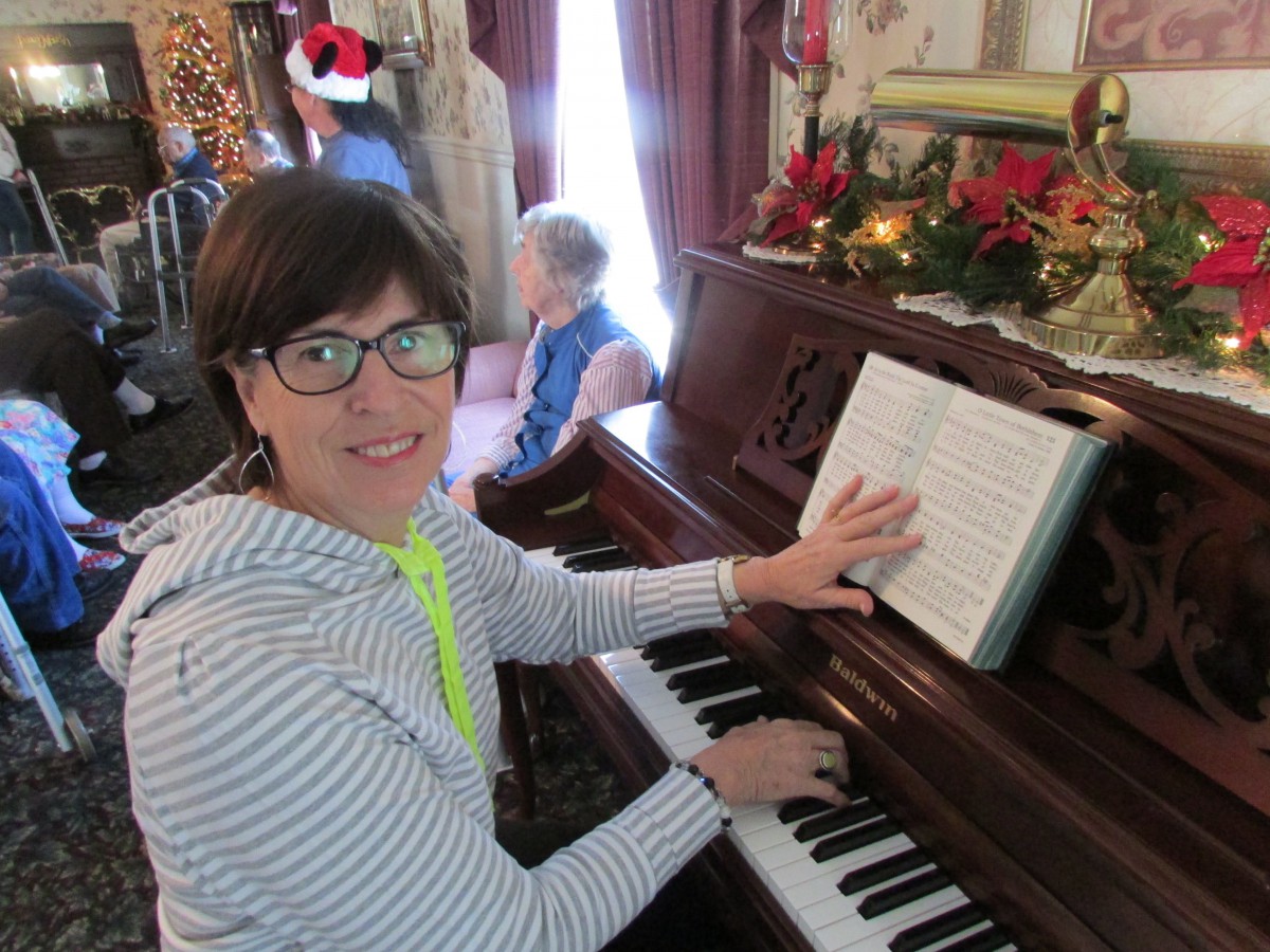 A woman wearing glasses plays the piano in a room decorated for the holidays.