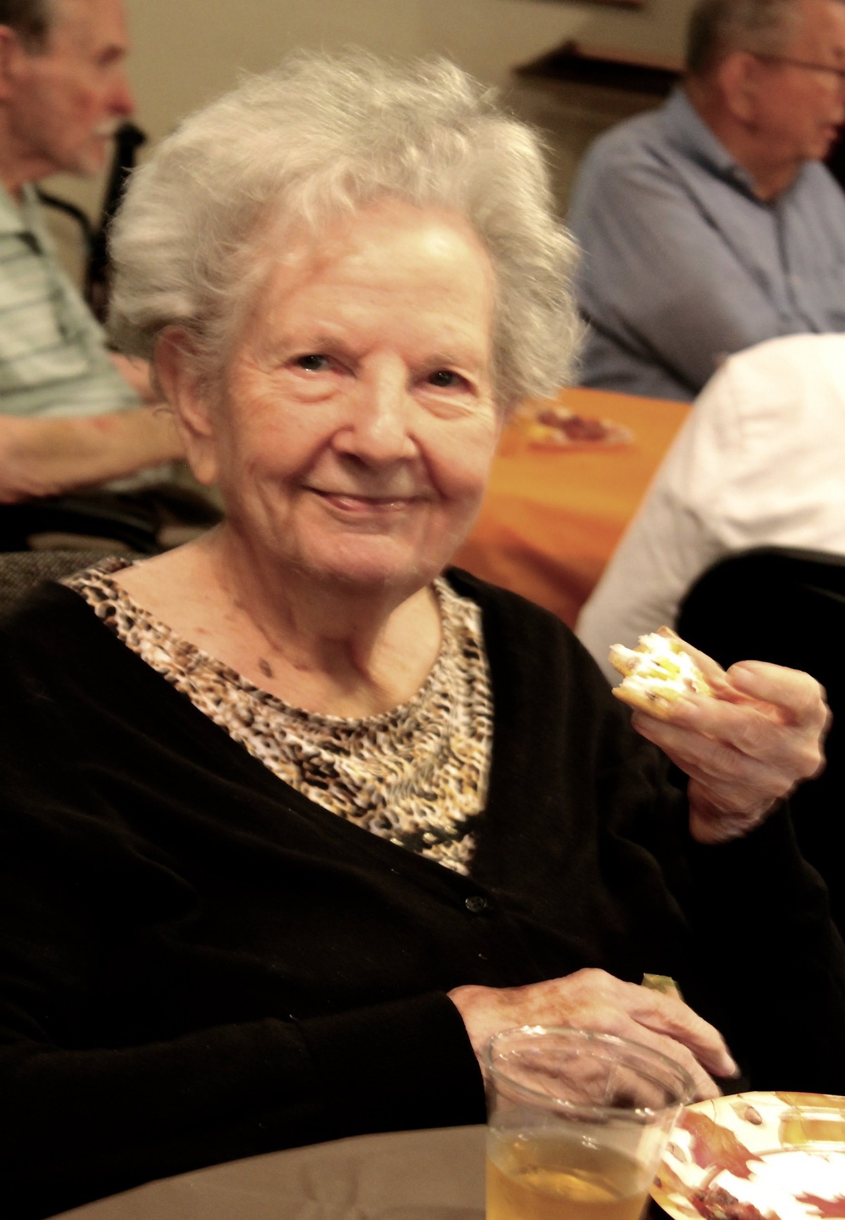 An elderly woman smiling while holding a piece of cake indoors.