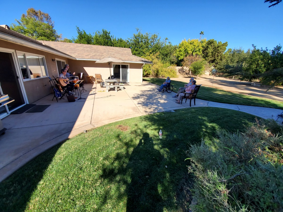A group of people enjoying a sunny day in the yard of a house with trees in the background.