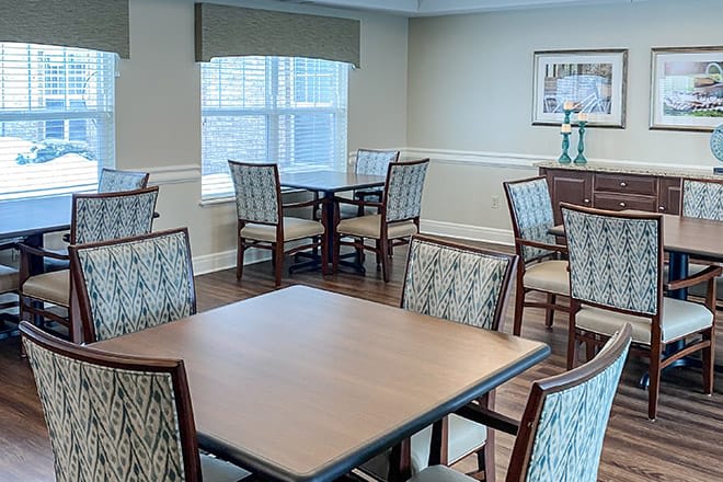 A well-lit dining area with multiple tables and chairs, featuring a window and hardwood flooring.