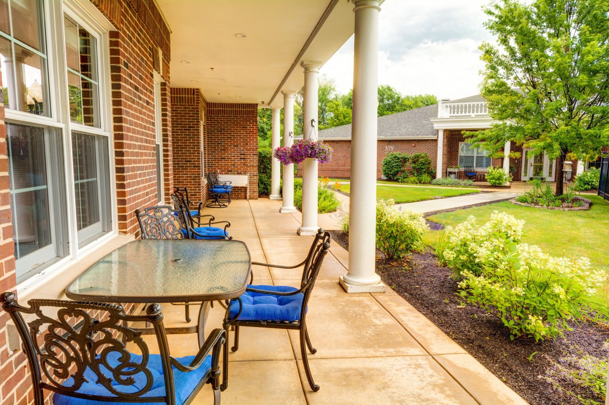 A cozy porch with a glass table and blue-cushioned chairs, overlooking a well-maintained garden.