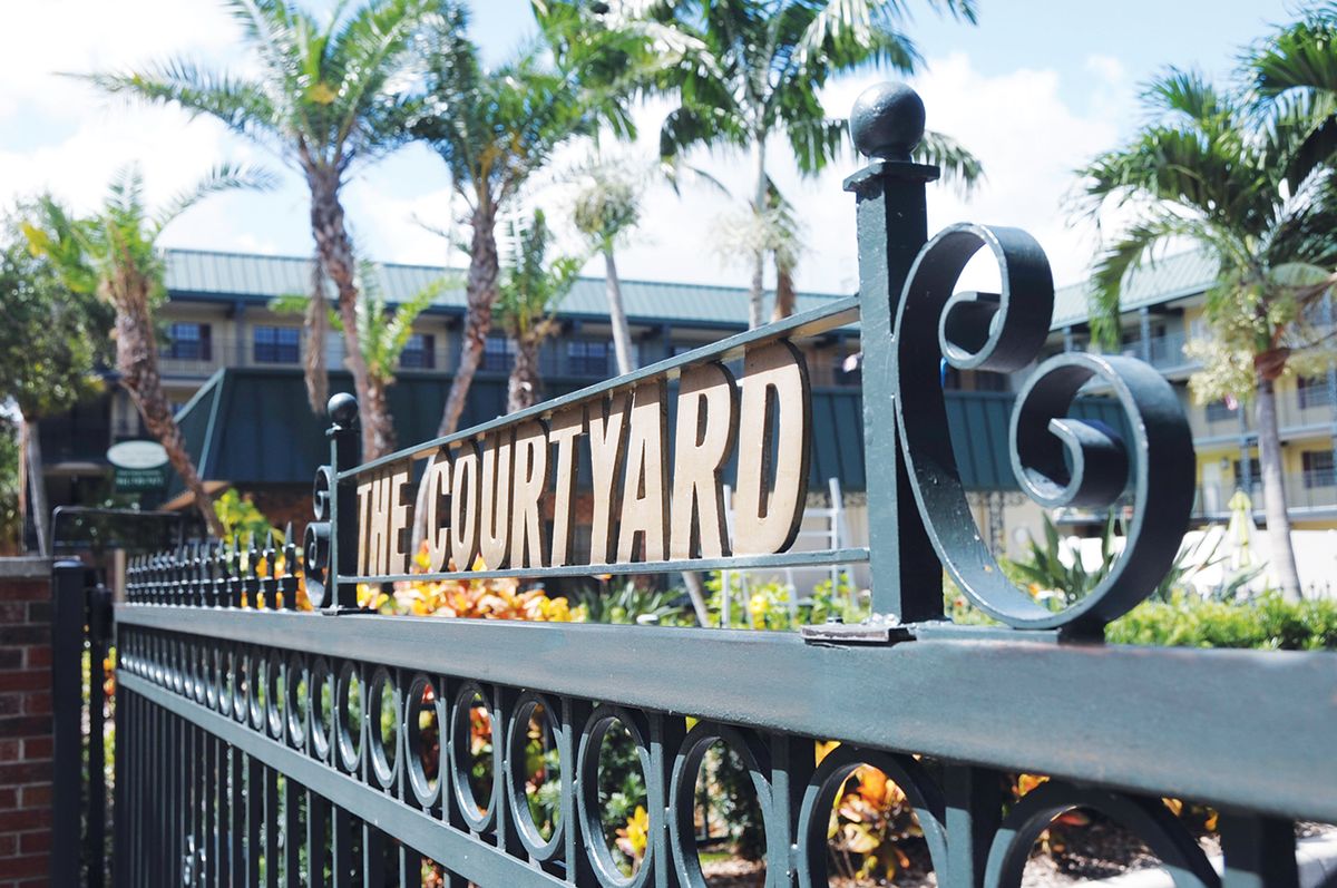 A decorative metal fence with a sign reading 'The Courtyard', set against a backdrop of palm trees and buildings.