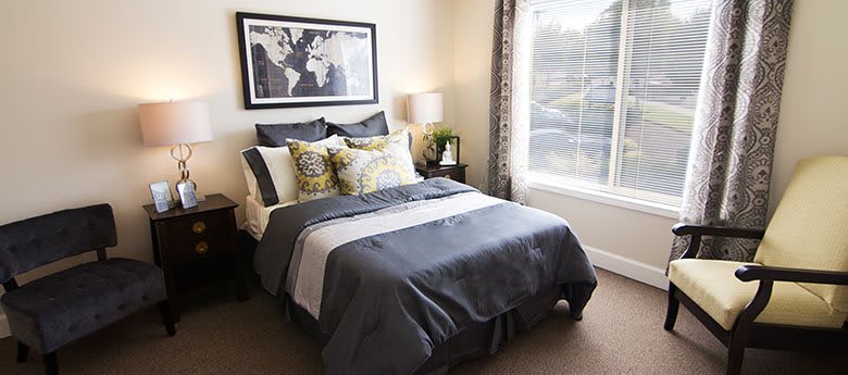A cozy and stylishly decorated bedroom featuring a neatly made bed with patterned pillows, a bedside lamp, and a large window.