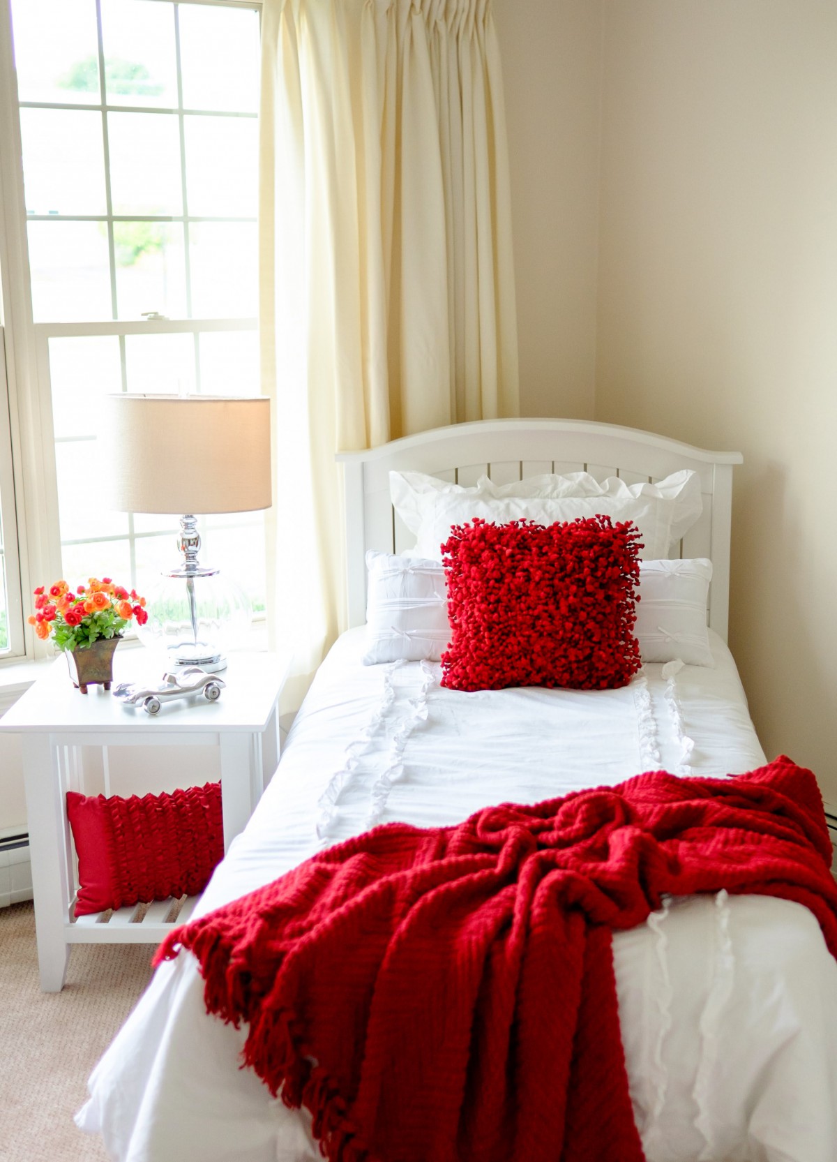 A cozy bedroom with white bedding and vibrant red accents, including a pillow and throw blanket. Natural light streams in through the window, illuminating the room.