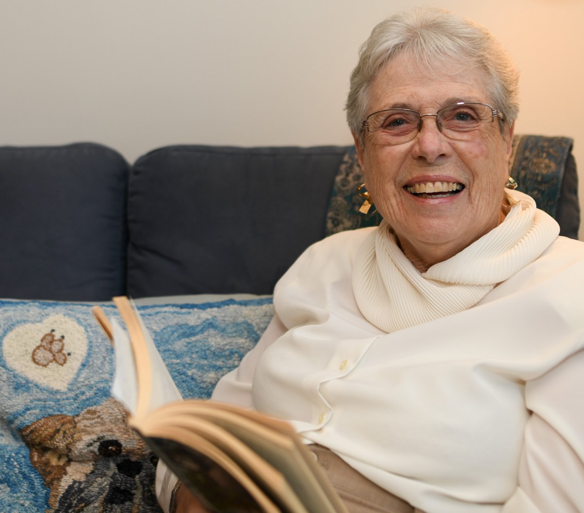 An elderly woman sitting on a couch, smiling while reading a book.