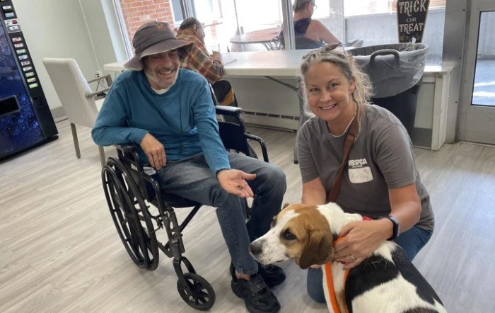 A man in a wheelchair interacts with a therapy dog and its handler indoors.