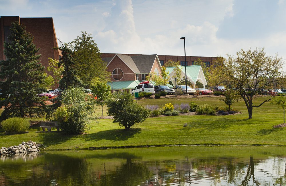 A scenic view of a building surrounded by lush greenery, with a small lake in the foreground and a cloudy sky overhead.