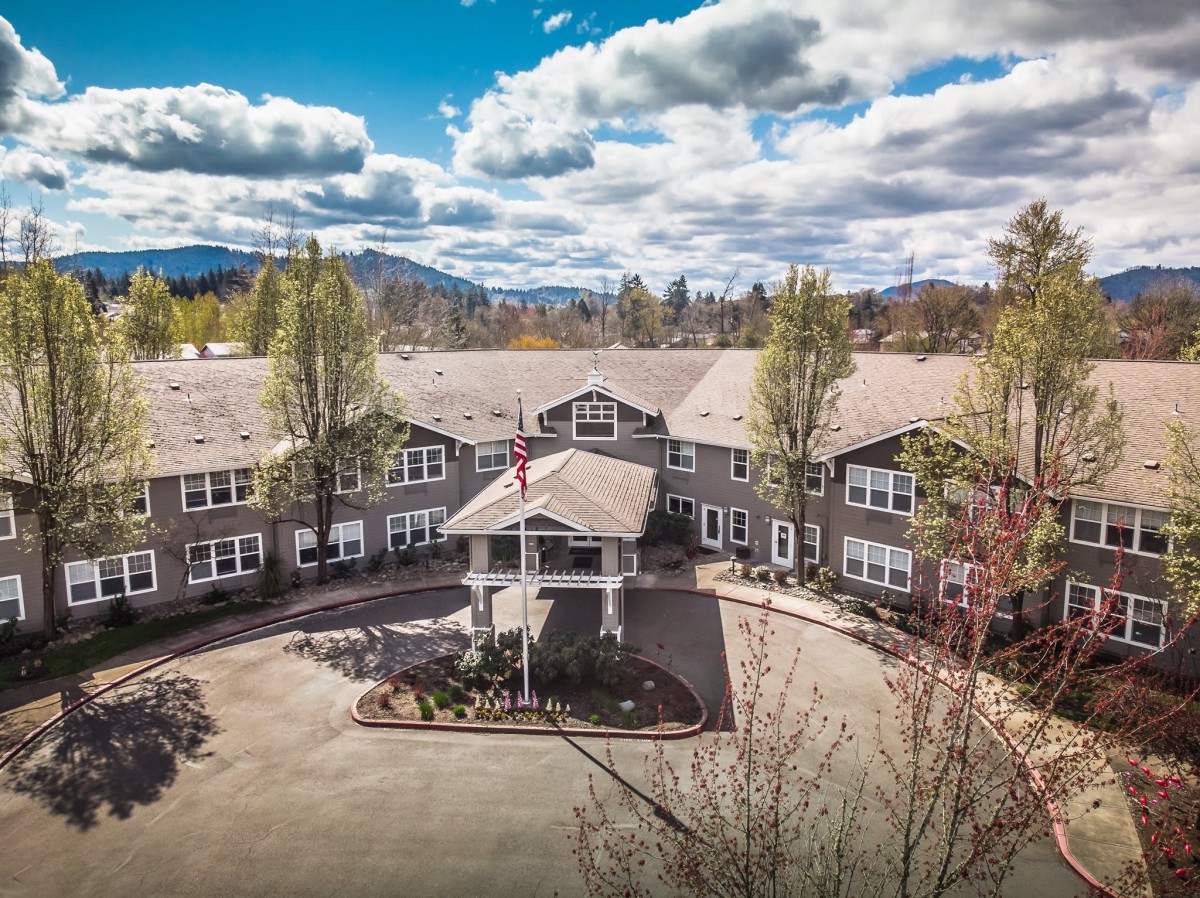 A large residential building surrounded by trees with a mountain backdrop under a partly cloudy sky.