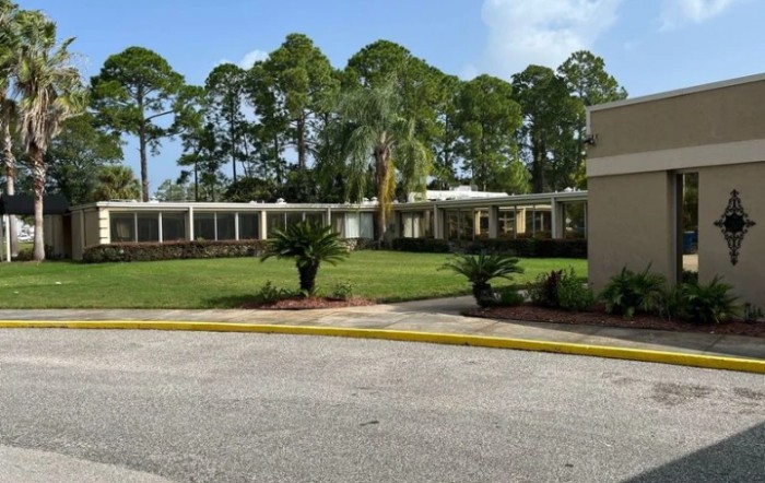 A low-rise building surrounded by lush greenery and palm trees, with a neat driveway in the foreground.