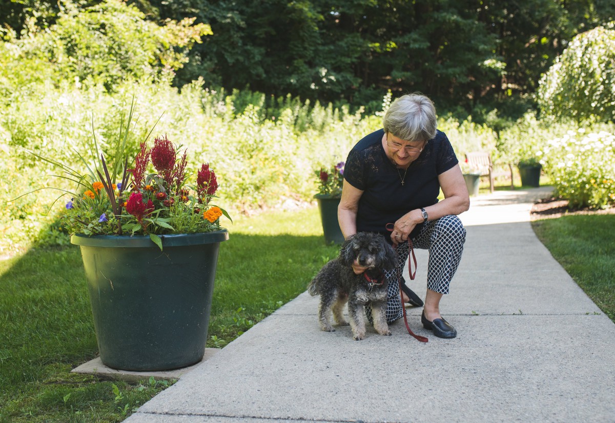 A woman kneels down to pet her dog in a garden setting with vibrant flowers.