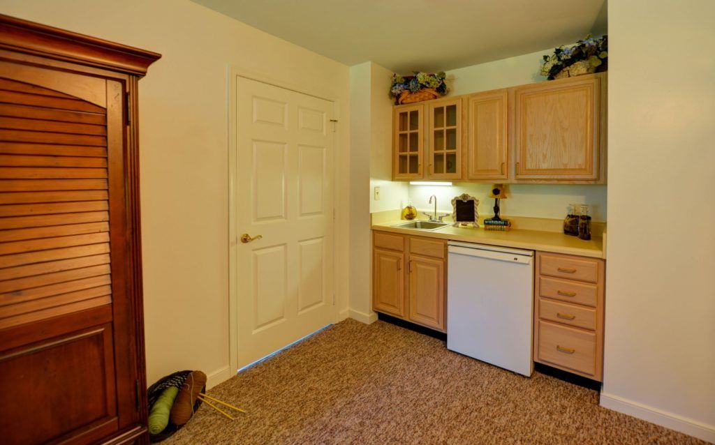 A small kitchen area with wooden cabinets, a compact refrigerator, and a carpeted floor.