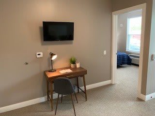 A cozy study area with a desk, chair, and lamp next to a wall-mounted TV, leading into a bedroom.