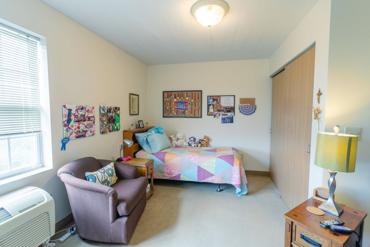 A cozy bedroom with a single bed covered in colorful bedding, decorated walls, and a comfortable chair.
