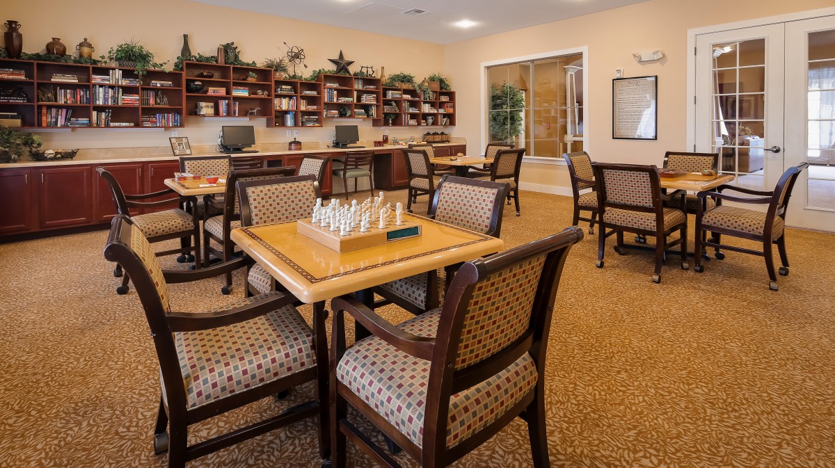 A cozy community room featuring a chessboard on a table, surrounded by chairs and bookshelves.