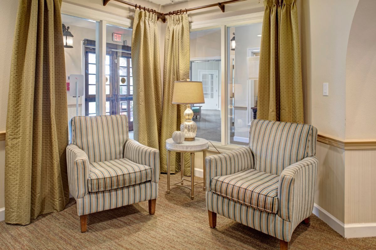 A cozy living room corner featuring two striped upholstered chairs, a small table with a lamp, and elegant curtains.
