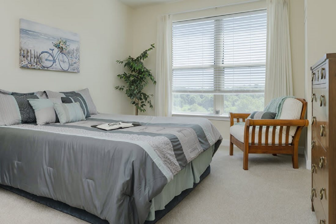 A cozy bedroom featuring a neatly made bed, a large window with shades, and a potted plant for decoration.