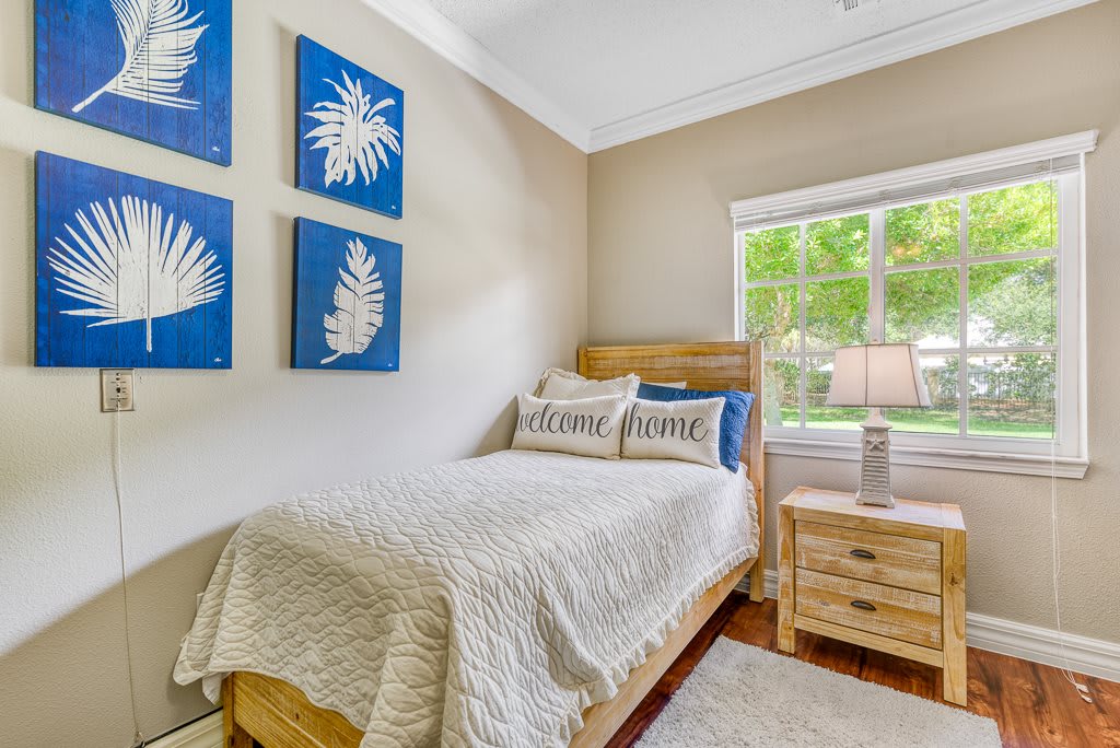 A cozy bedroom with a single bed and decorative pillows, featuring blue leaf-themed wall art and a wooden dresser by the window.
