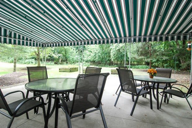 A patio with tables and chairs under a striped awning overlooking a garden.