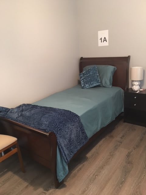 A neatly made single bed in a small bedroom with blue bedding and a nightstand with a lamp.