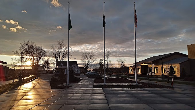 Three flagpoles stand silhouetted against a cloudy sunset sky near buildings.