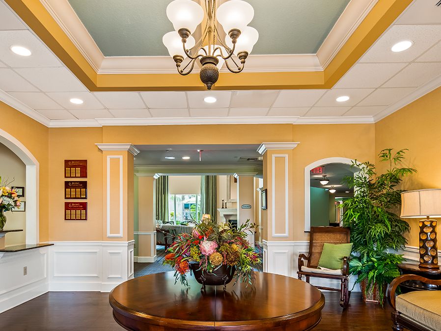 A warmly lit lobby with a central table featuring a floral arrangement and an elegant chandelier overhead.