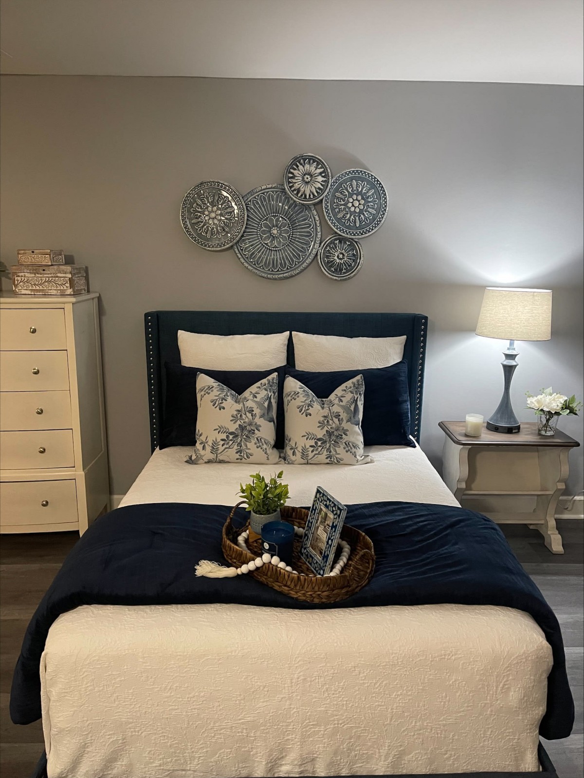 A cozy, well-decorated bedroom featuring a bed with decorative pillows and a blanket, a nightstand with a lamp, and wall art.