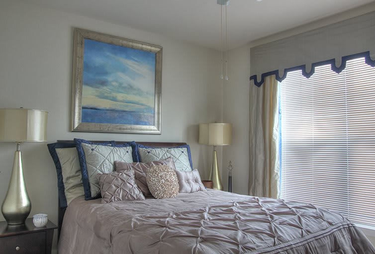 A cozy bedroom with a neatly made bed, featuring decorative pillows and a large painting above the headboard. A lamp stands beside the bed near a window with drapes.